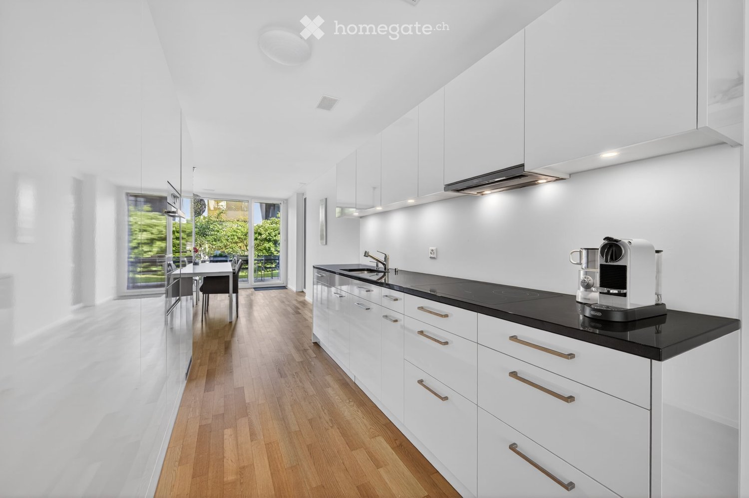 Modern kitchen with white cabinets, black countertops, and a coffee machine on the counter. The space features a dining area with a table and chairs, and large windows offering a view of greenery outside.