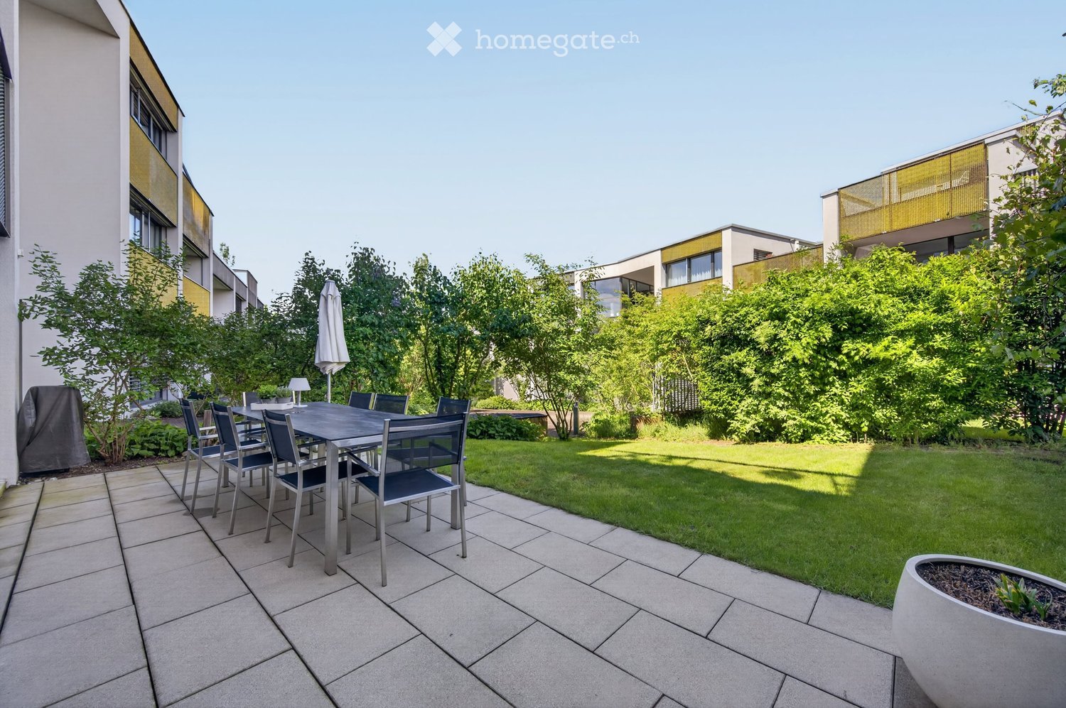 Garden area with a tiled patio, a dining table with chairs, an umbrella, and a well-maintained lawn surrounded by trees and modern buildings.
