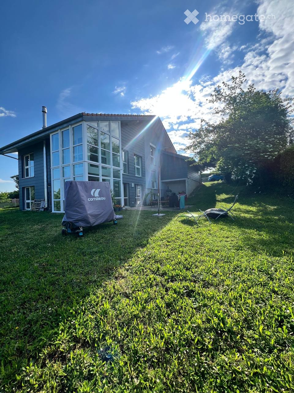 A modern house with large windows and a covered patio area, surrounded by a well-maintained lawn and outdoor furniture.