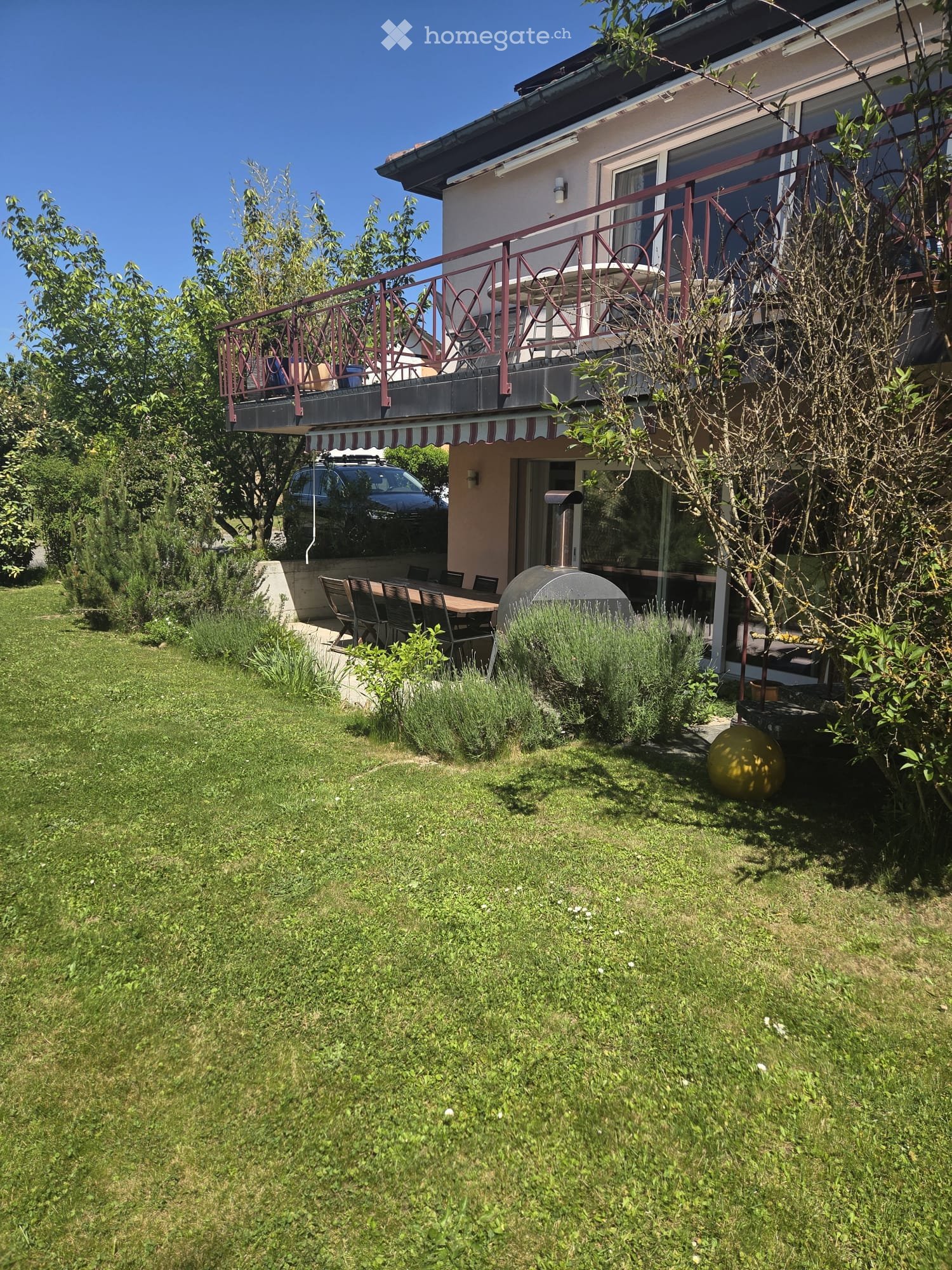 Lush garden with a well-maintained lawn, a patio area with a table and chairs, a round outdoor ball, and a balcony with a railing and outdoor furniture.