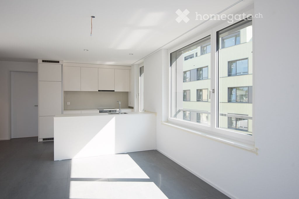 Modern, minimalist kitchen with white cabinets, a central island, and large windows offering a view of neighboring buildings.