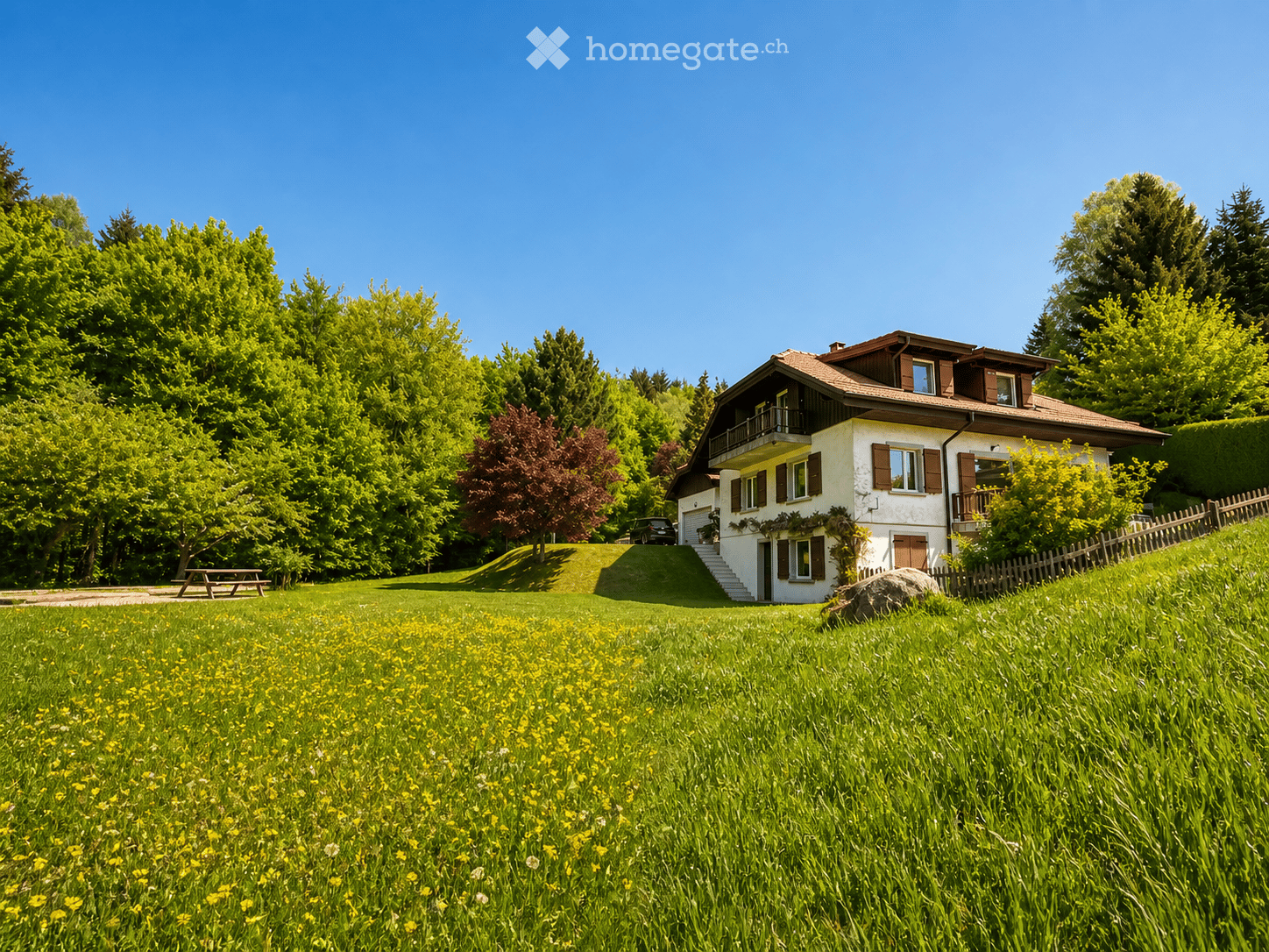 A two-story house with a red-tiled roof, white walls, and brown shutters, situated on a raised ground floor with a grassy lawn and wildflowers in the foreground.