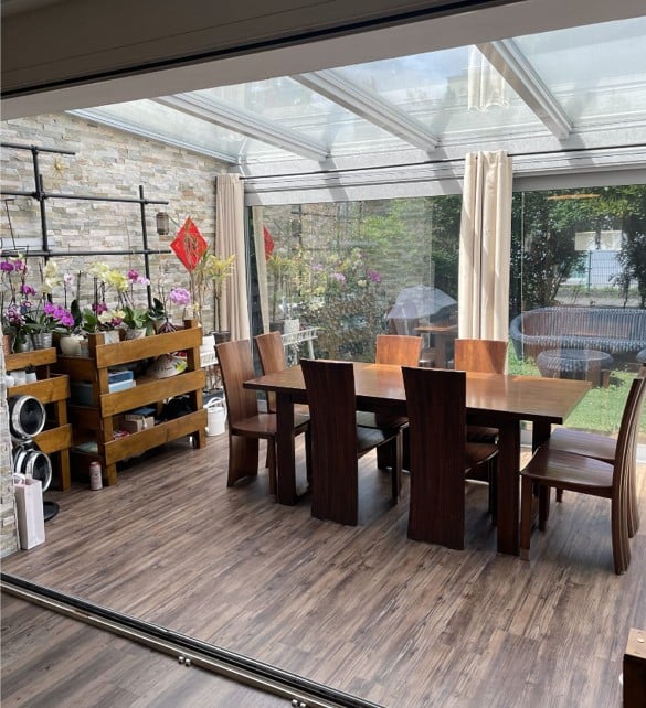 Bright, airy dining area with wooden flooring, large glass ceiling, and a view of a garden. Features a wooden dining table with six chairs, shelves with flower arrangements, and a curtained window.
