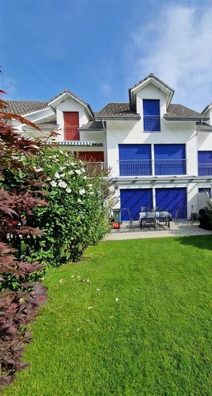 Modern two-story house with white exterior, blue shutters, and a well-maintained garden with a patio seating area.
