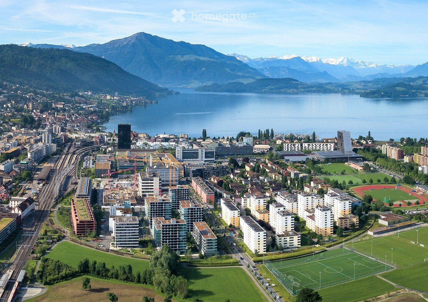 Aerial view of a cityscape with modern apartment buildings, green spaces, a lake, and surrounding mountains.