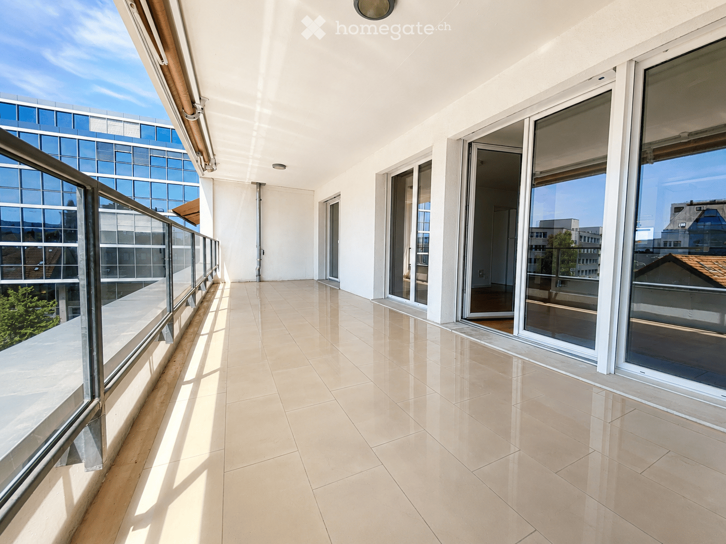 Spacious balcony with light-colored tiled flooring, large glass sliding doors, and a modern glass railing, offering views of surrounding buildings and greenery.