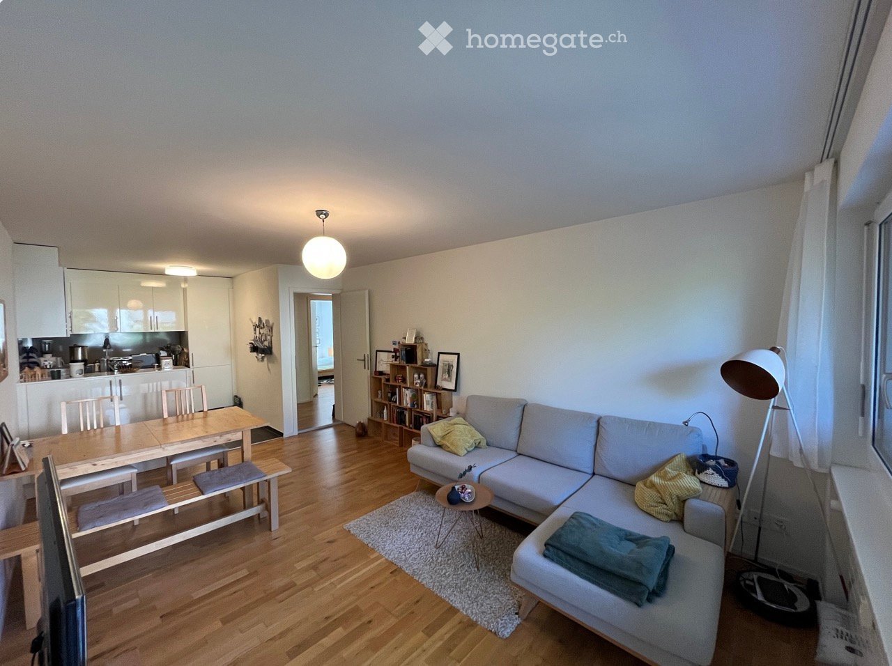 Bright, open-plan living and dining area with wooden flooring, a grey sectional sofa, a small coffee table, a dining table with chairs, a bookshelf, and a modern kitchen area with cabinets and appliances.