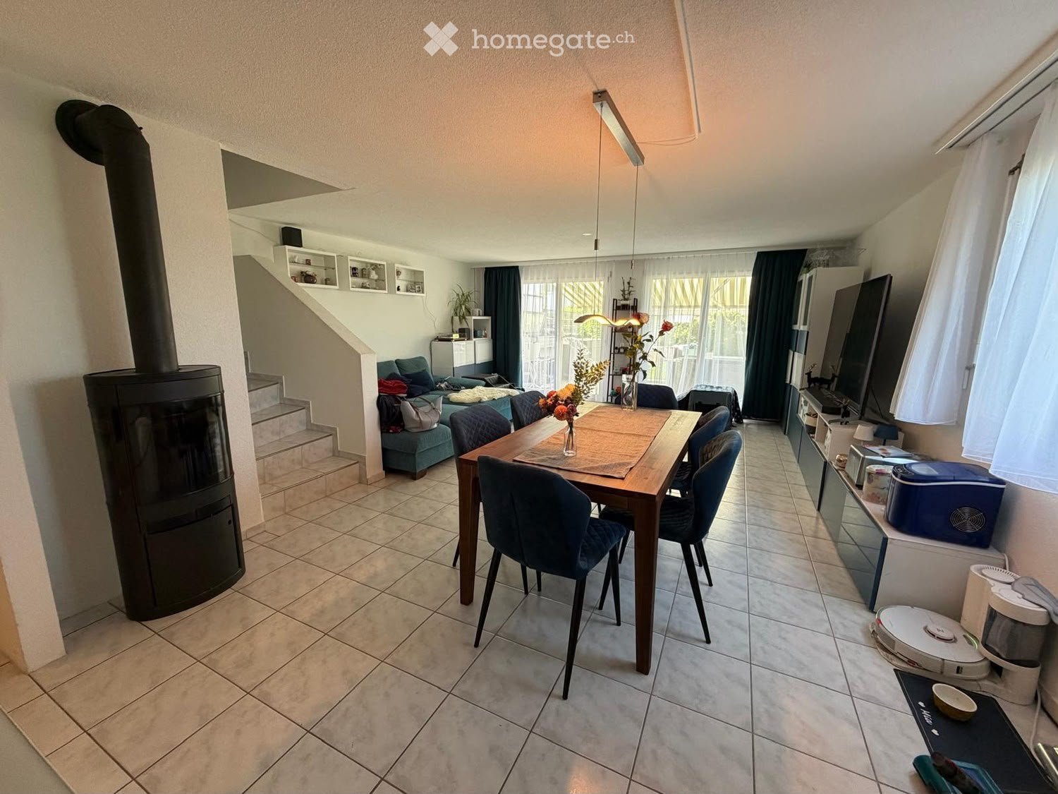 A well-lit living and dining area with a tiled floor, a large wooden dining table with chairs, a sofa, a fireplace, and a television. There are also shelves and a staircase leading to another level.