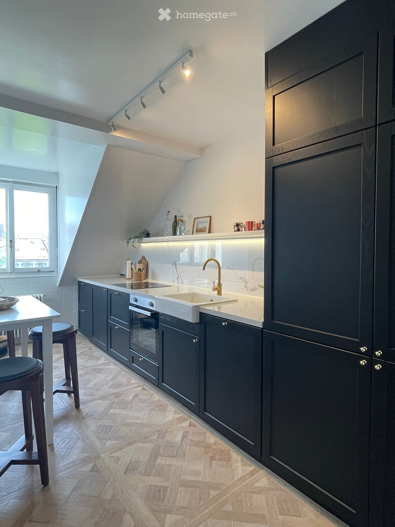 Modern kitchen with dark cabinets, white countertops, and a built-in sink and oven. Features include a sleek faucet, a small dining area with a table and stools, and decorative items on the countertop.