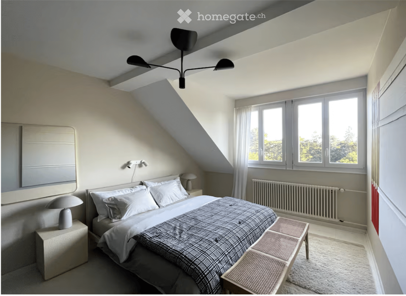 Cozy attic bedroom with slanted ceilings, large windows, a neatly made bed, nightstands with lamps, a small stool, and a mirror on the wall.