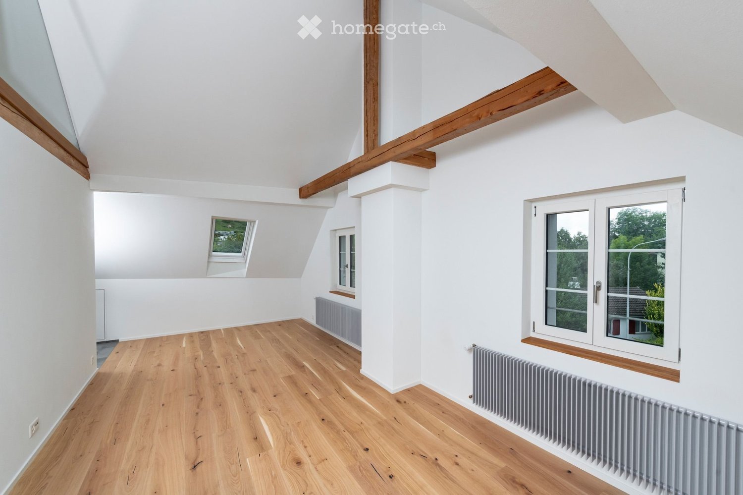 Bright, empty attic room with wooden flooring, white walls, and exposed wooden beams. Features multiple windows, including a skylight, and built-in radiators.