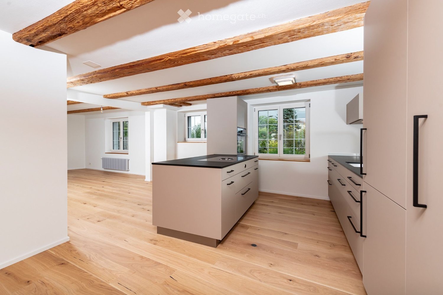 Modern kitchen with light-colored cabinets, a central island, and wooden flooring. Features include a stove, sink, and ample natural light from large windows.