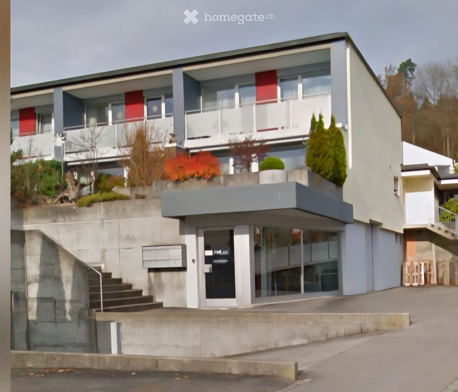 Modern two-story building with white and grey exterior, large windows, balconies, and landscaped garden area with plants and trees.
