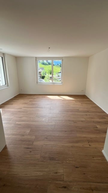 Empty room with wooden flooring, white walls, and large windows offering a scenic view of greenery outside.