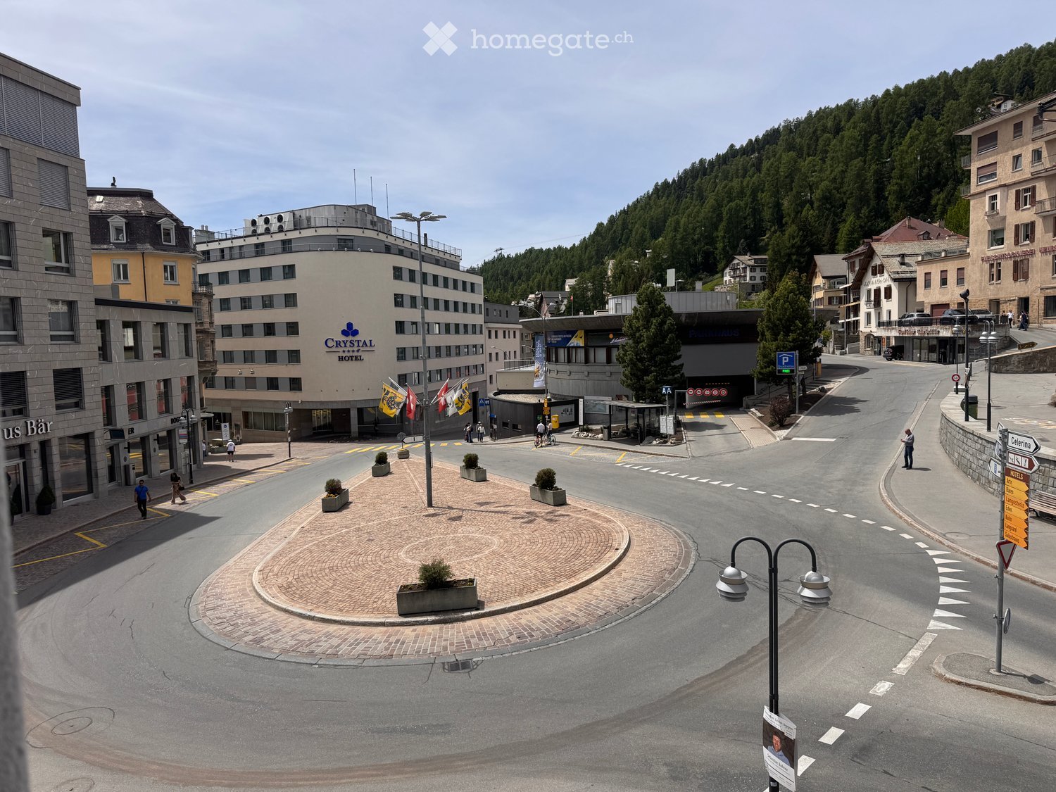 A town square with a circular brick-paved area, surrounded by modern buildings including a hotel, with a backdrop of forested hills.