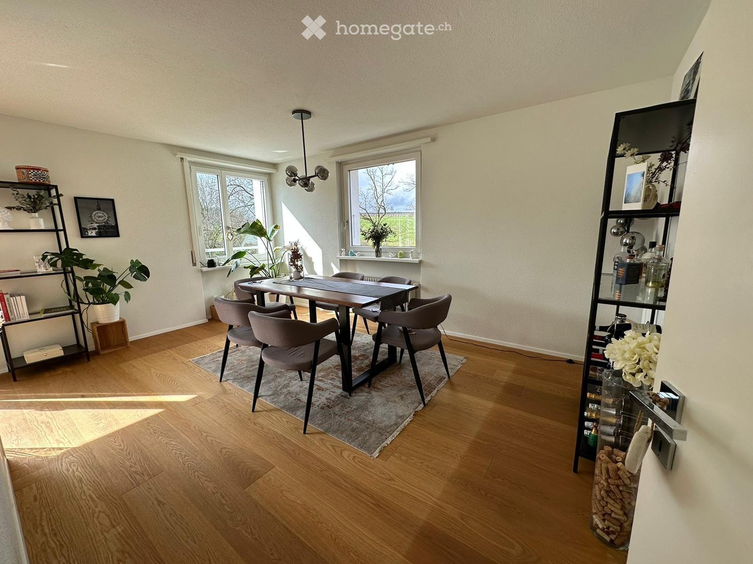 Dining area with a wooden dining table, six chairs, shelves with books and plants, and a wine rack in the corner.