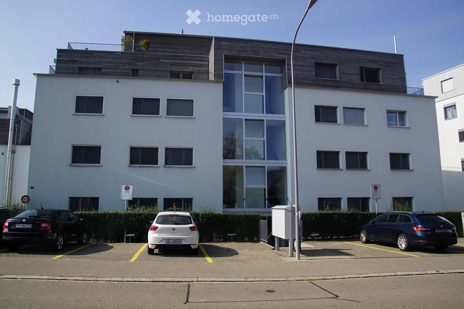Modern multi-story building with large windows and a flat roof, surrounded by a parking area with several cars.