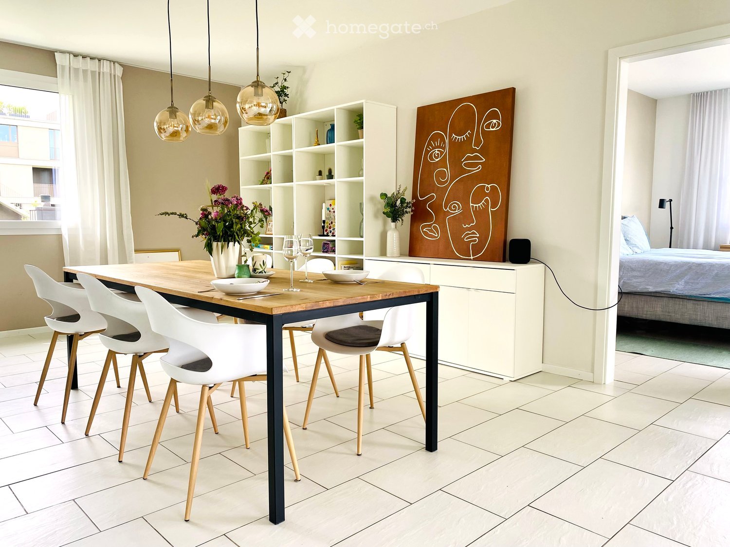 Modern dining area with a wooden table, white chairs, and a shelf with decorative items. A large abstract art piece is mounted on the wall, and a vase with flowers sits on the table.
