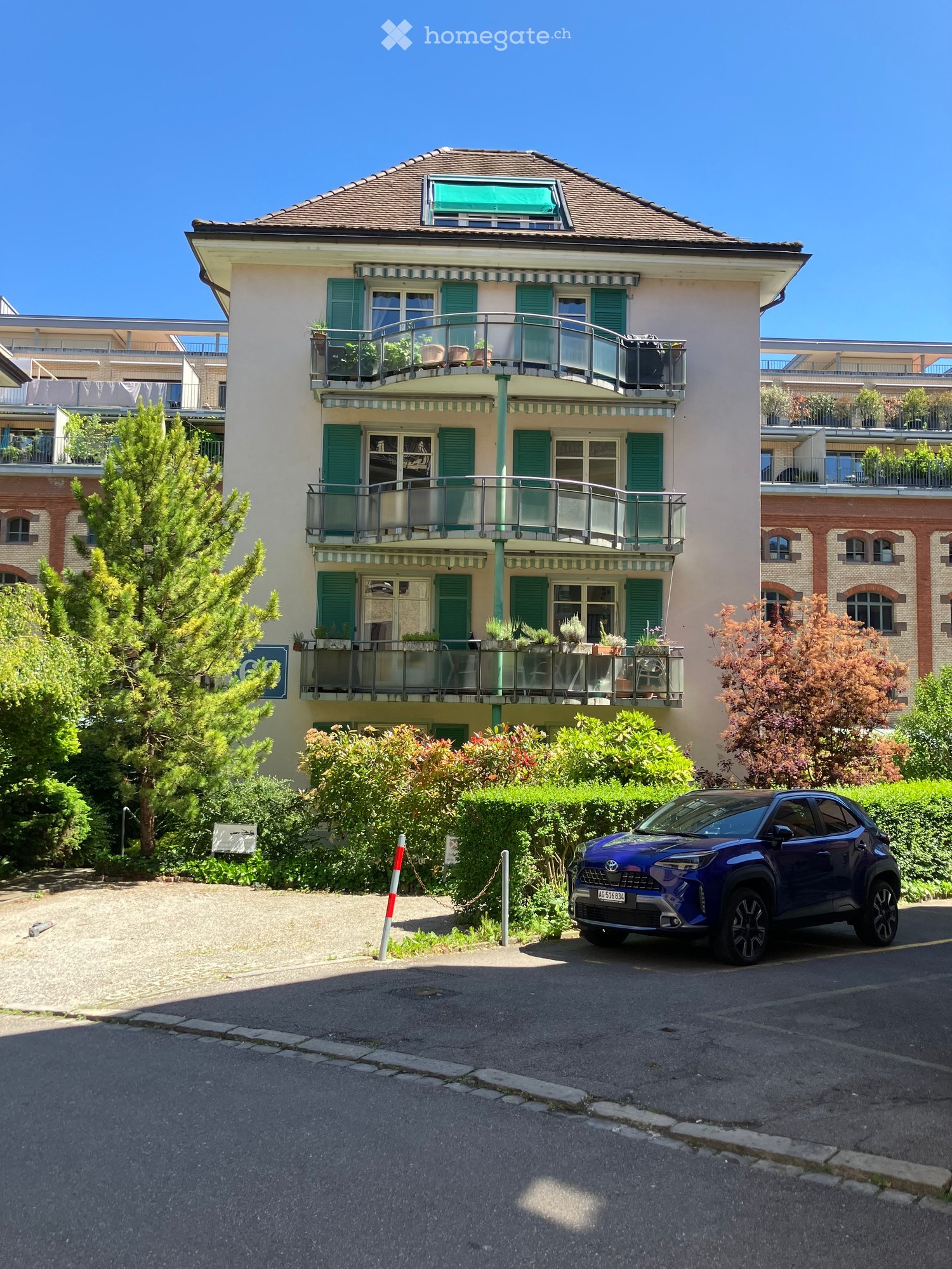 A multi-story residential building with green shutters, balconies, and potted plants. A blue car is parked in front.