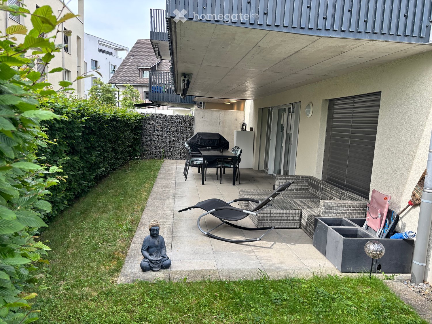 Small outdoor patio area with a table, chairs, sun lounger, and a Buddha statue, surrounded by greenery and a modern building.