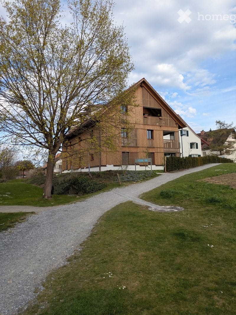 A two-story wooden house with a sloped roof, large windows, and a balcony, surrounded by a well-maintained lawn and gravel pathway.