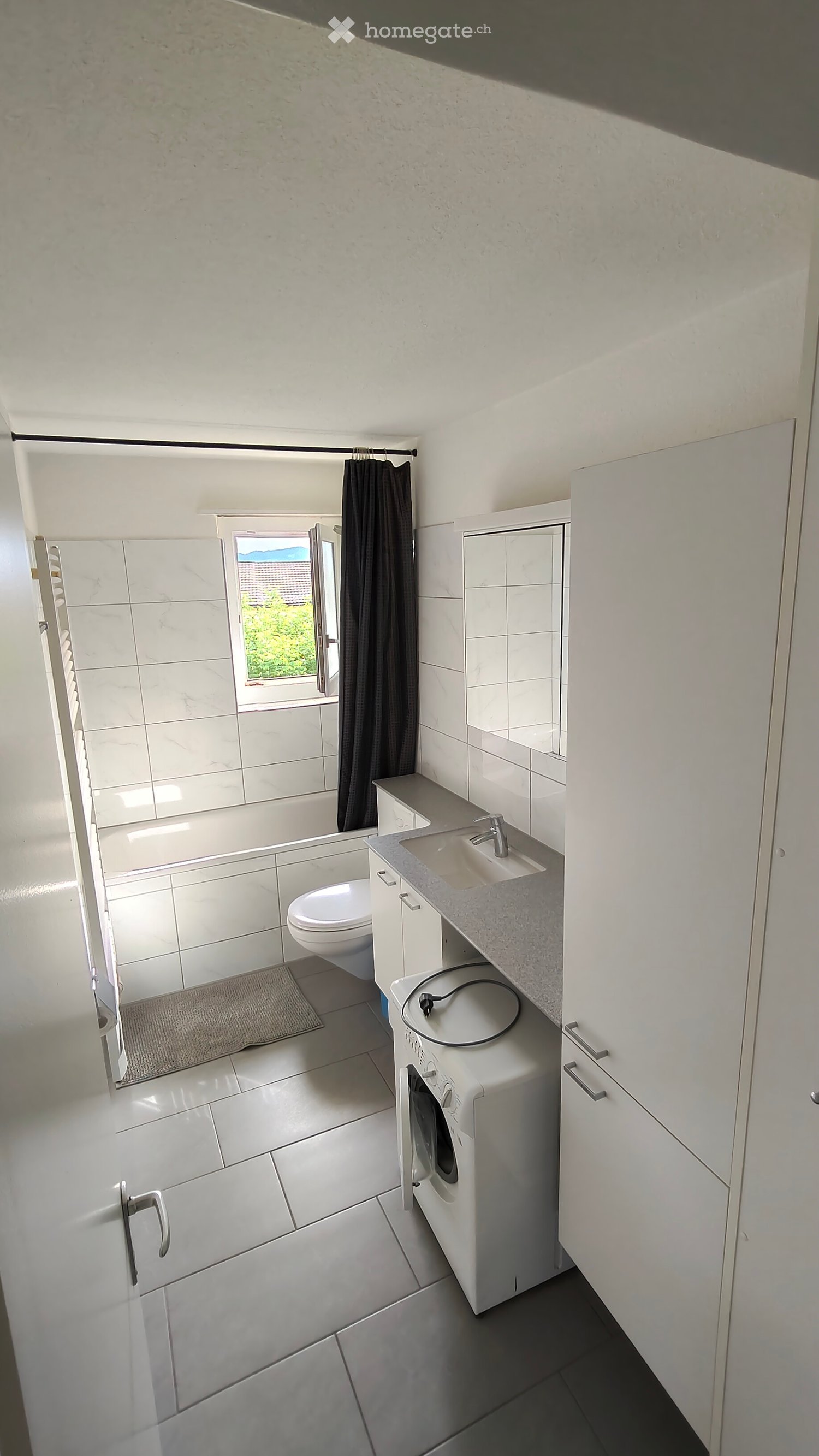 Modern bathroom with white tiles, a sink, toilet, bathtub, washing machine, and a window with a curtain.