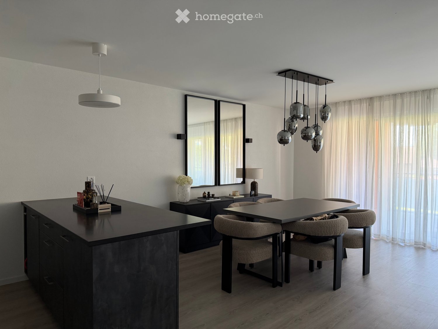 Modern dining area with a sleek black dining table, four chairs, a large kitchen island, and contemporary lighting fixtures.