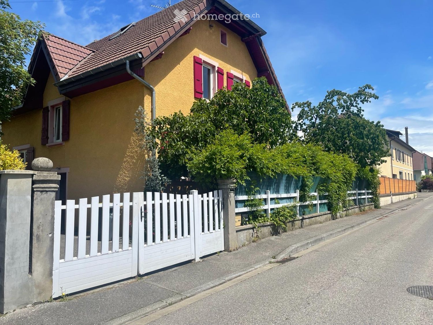 Two-story yellow house with red shutters, surrounded by greenery and a white picket fence.