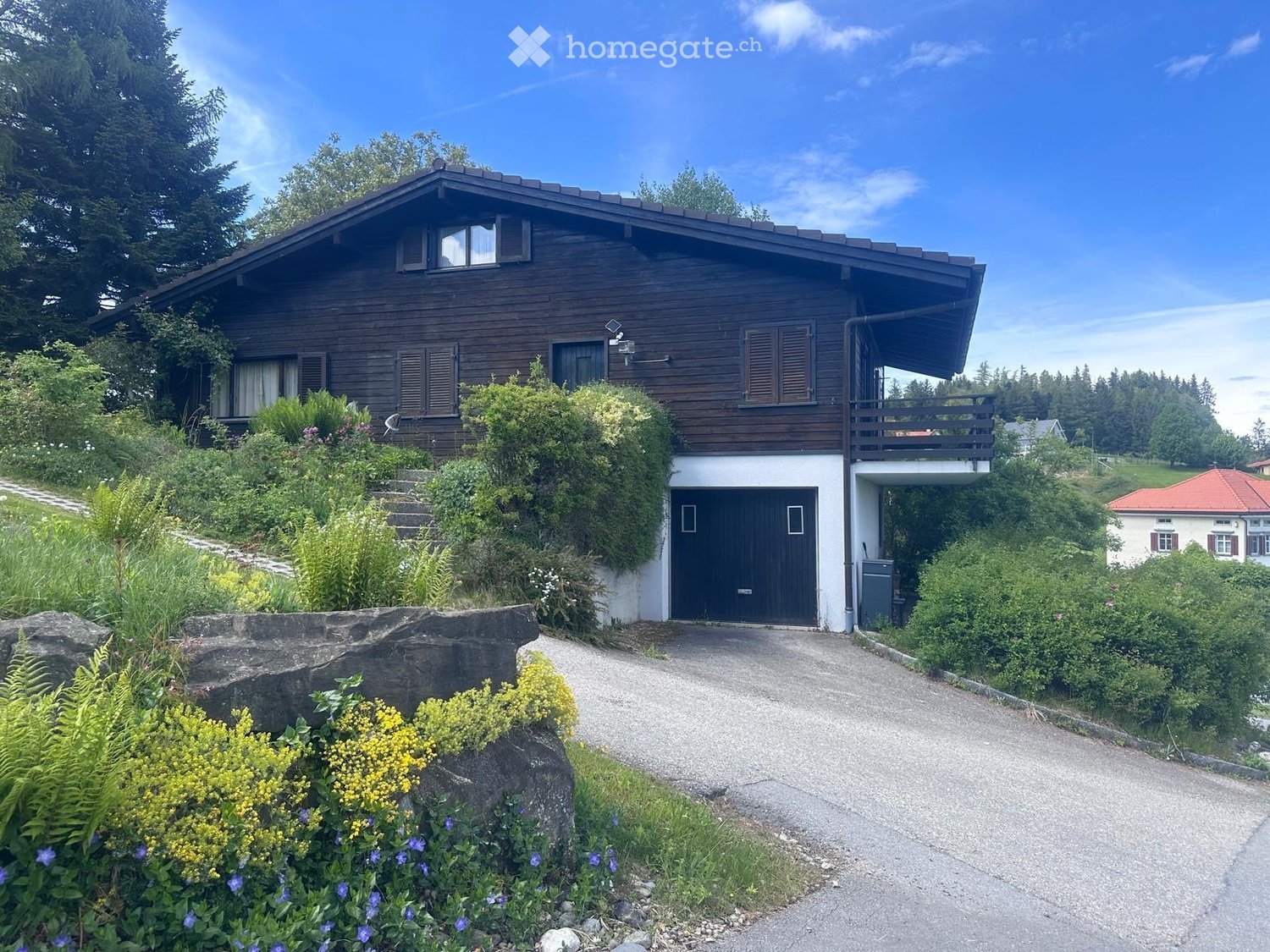 Two-story wooden house with a garage, surrounded by greenery and a driveway.