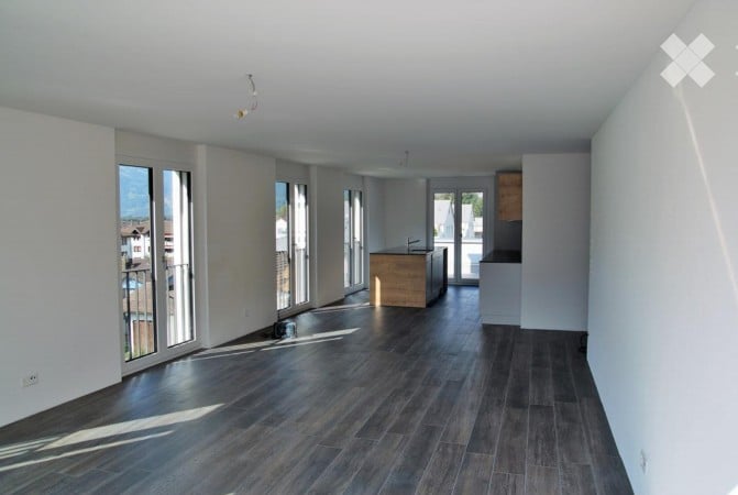 Empty room with dark wooden flooring, white walls, large windows, and a small kitchen area visible in the background.
