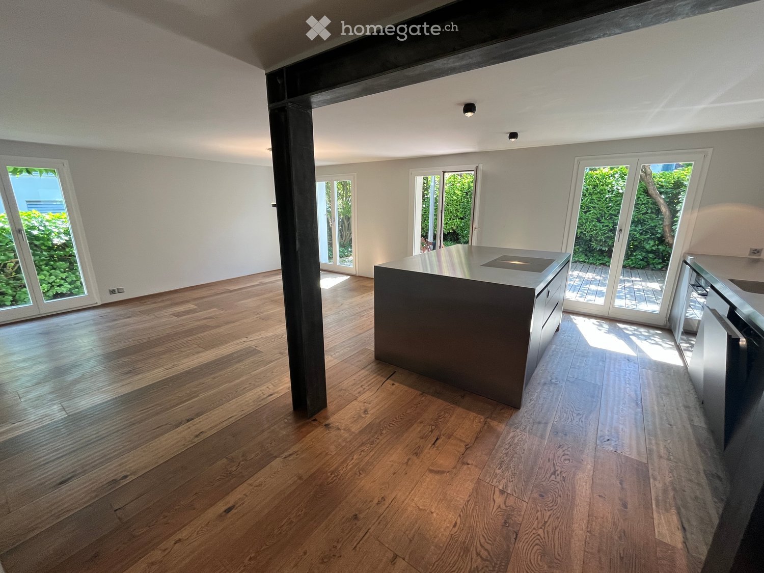 Open-plan space with wooden flooring, large windows, and a central island with a built-in stove. The room is empty, with no furniture or appliances visible.