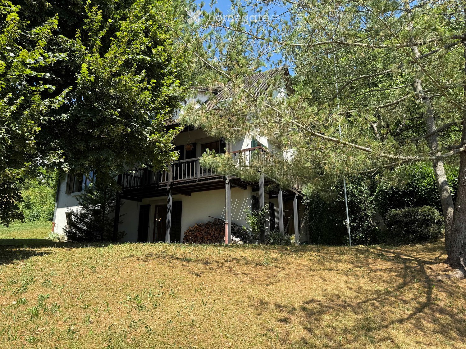 Two-story house with white exterior, multiple windows, and a balcony on the upper level, surrounded by trees and greenery.