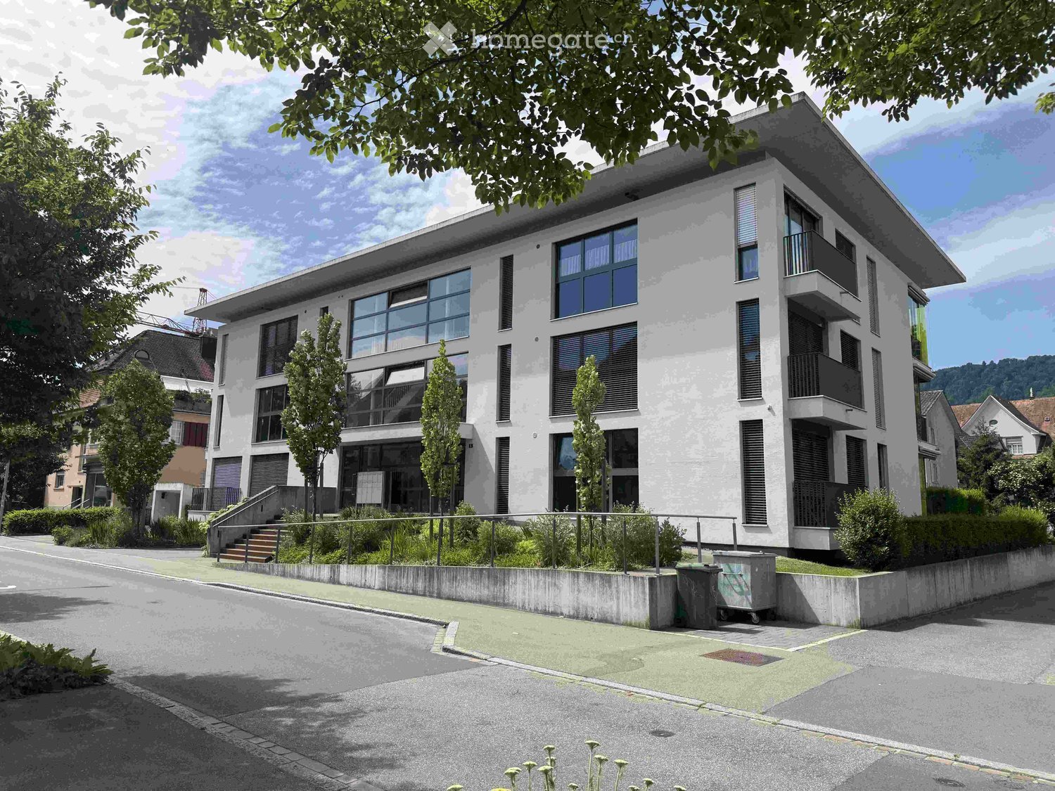 Modern multi-story residential building with white facade, large windows, and balconies. The building is surrounded by greenery and has a paved entrance area.