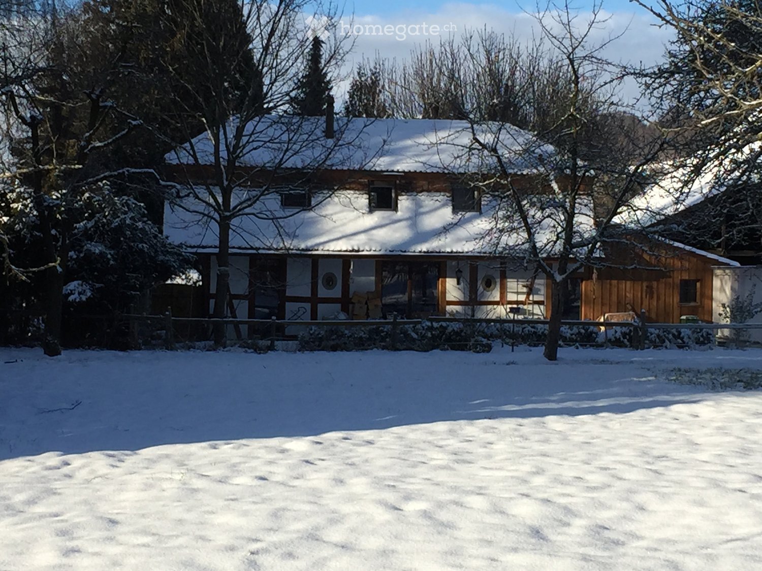 a detached house with snow on the roof, wooden exterior, several windows, a fenced garden, and surrounded by trees