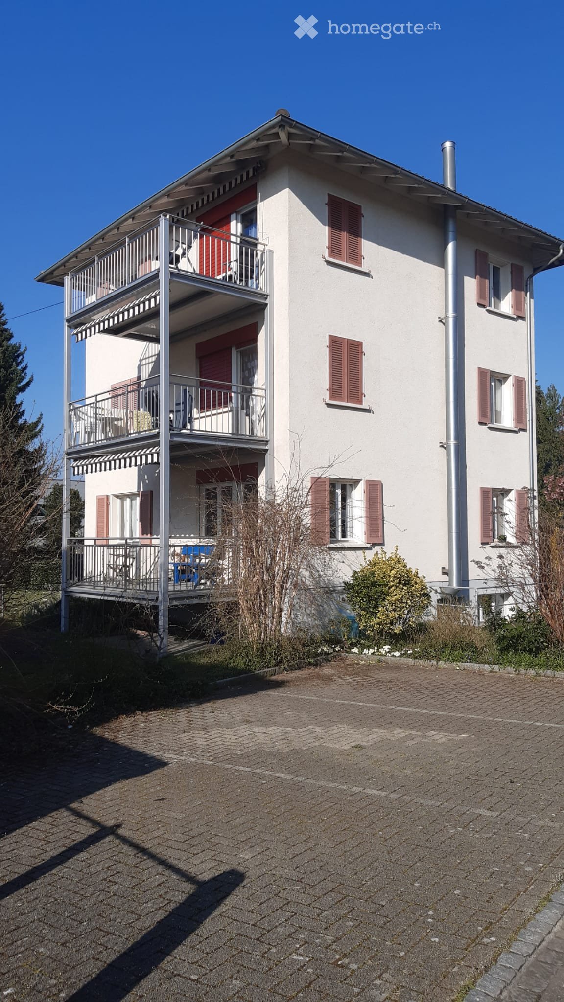 Two-story house with white walls, red shutters, and a balcony with metal railings. The house has multiple windows and a brick driveway.