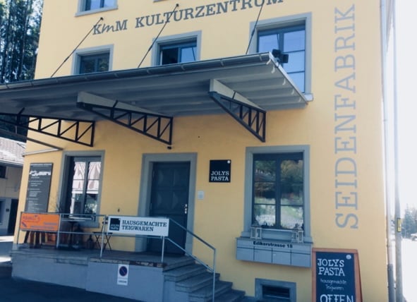 Yellow building with signage reading "KÜmm Kulturzentrum" and "Seidenfabrik". Features a covered entrance, steps, and a small signboard advertising "Joey's Pasta".