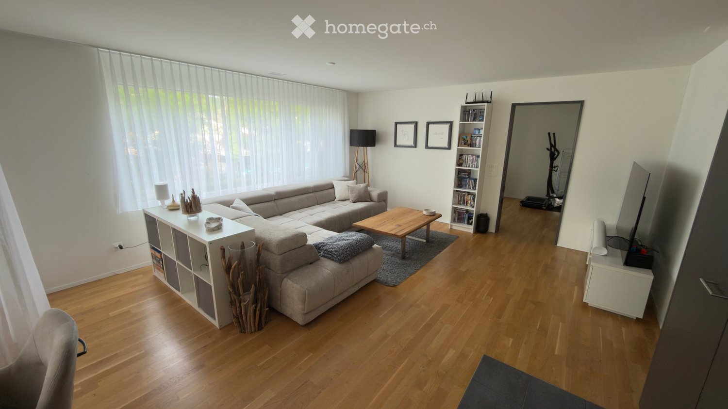 Modern living room with light wooden flooring, large windows with sheer curtains, grey sectional sofa, wooden coffee table, bookshelf, floor lamp, and exercise bike in the background.