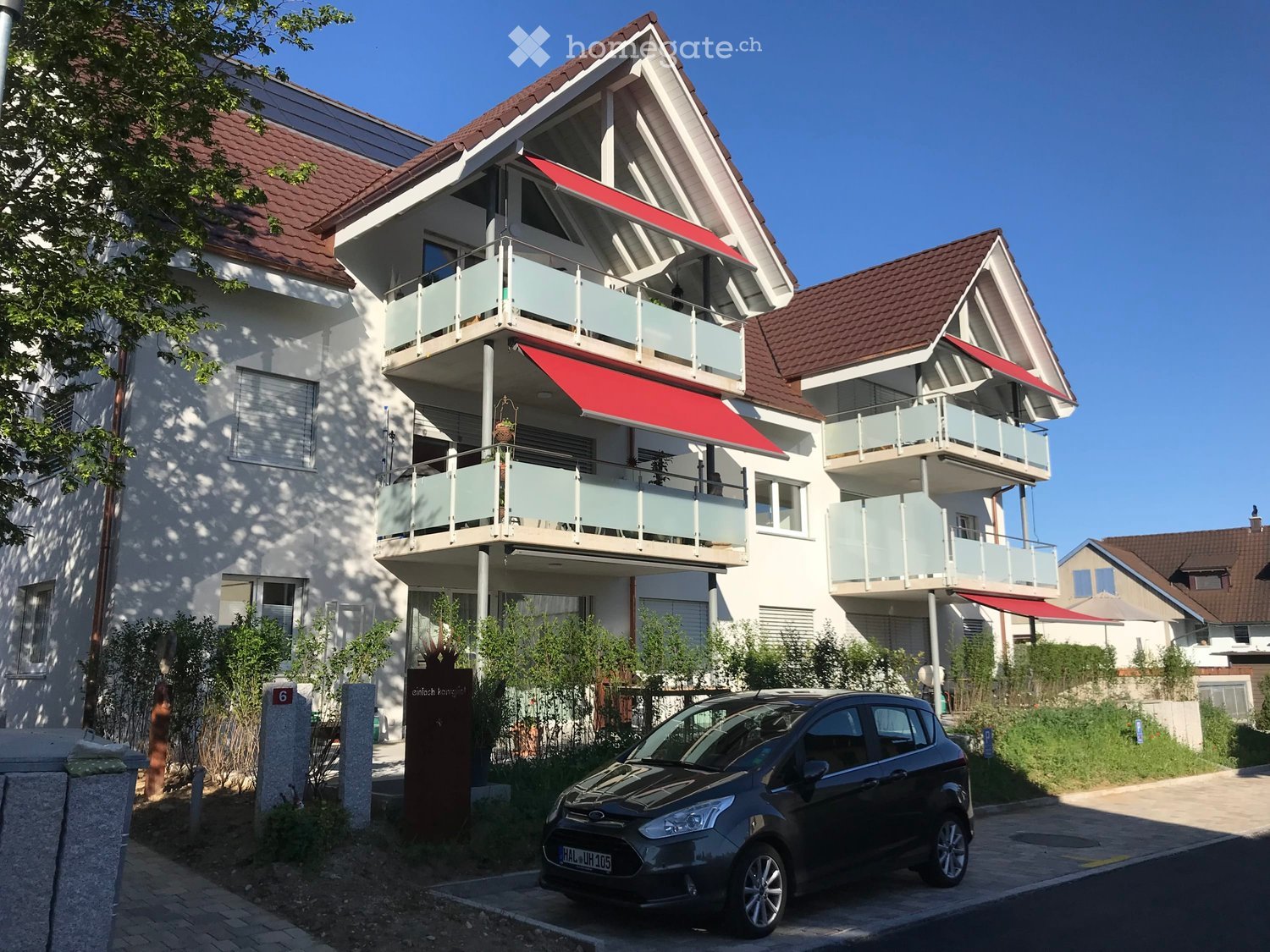 Two-story residential building with white walls, red-tiled roof, and balconies with red awnings. A car is parked in front.