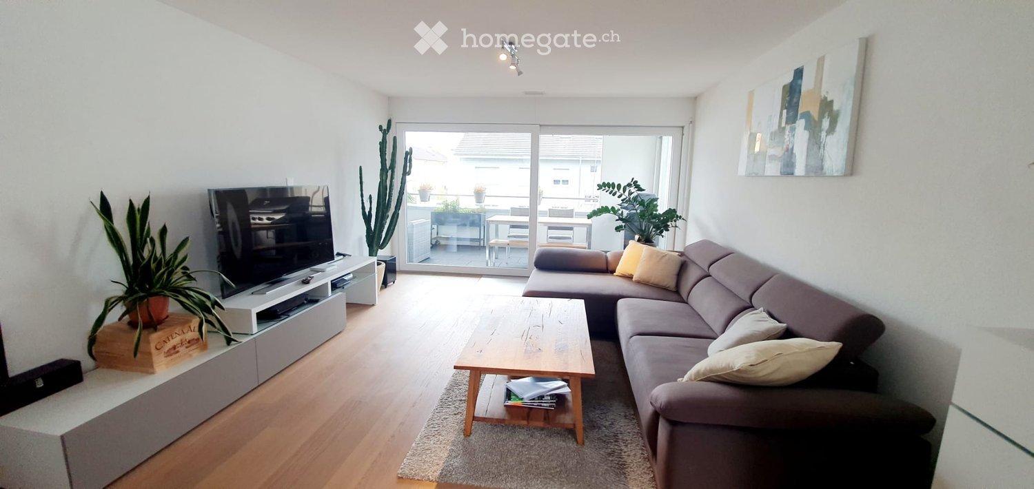 Modern living room with a grey sectional sofa, wooden coffee table, flat-screen TV, and large sliding glass doors leading to a balcony.