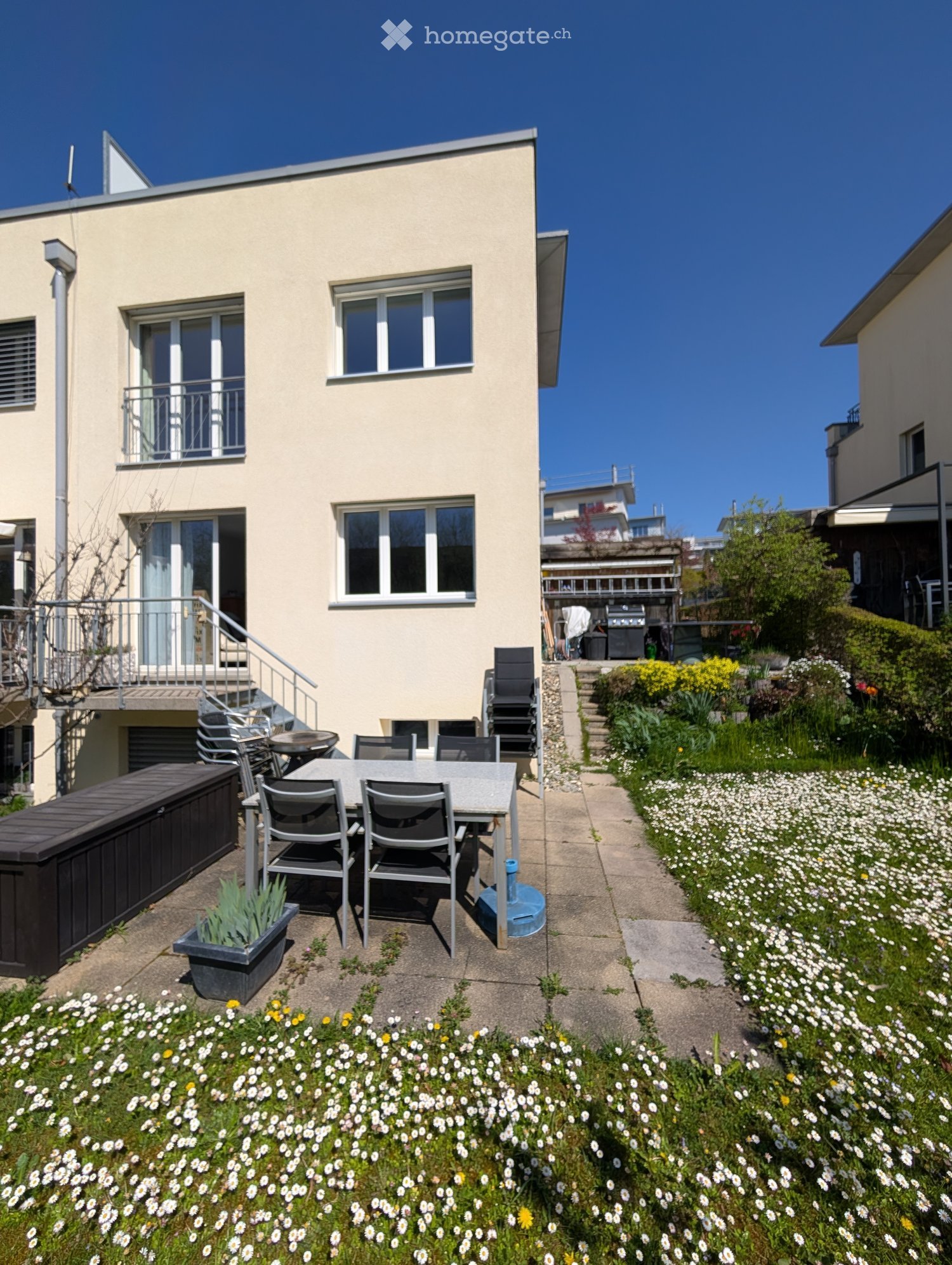 Garden area with a patio table and chairs, surrounded by white and yellow flowers, leading to a house with a balcony.