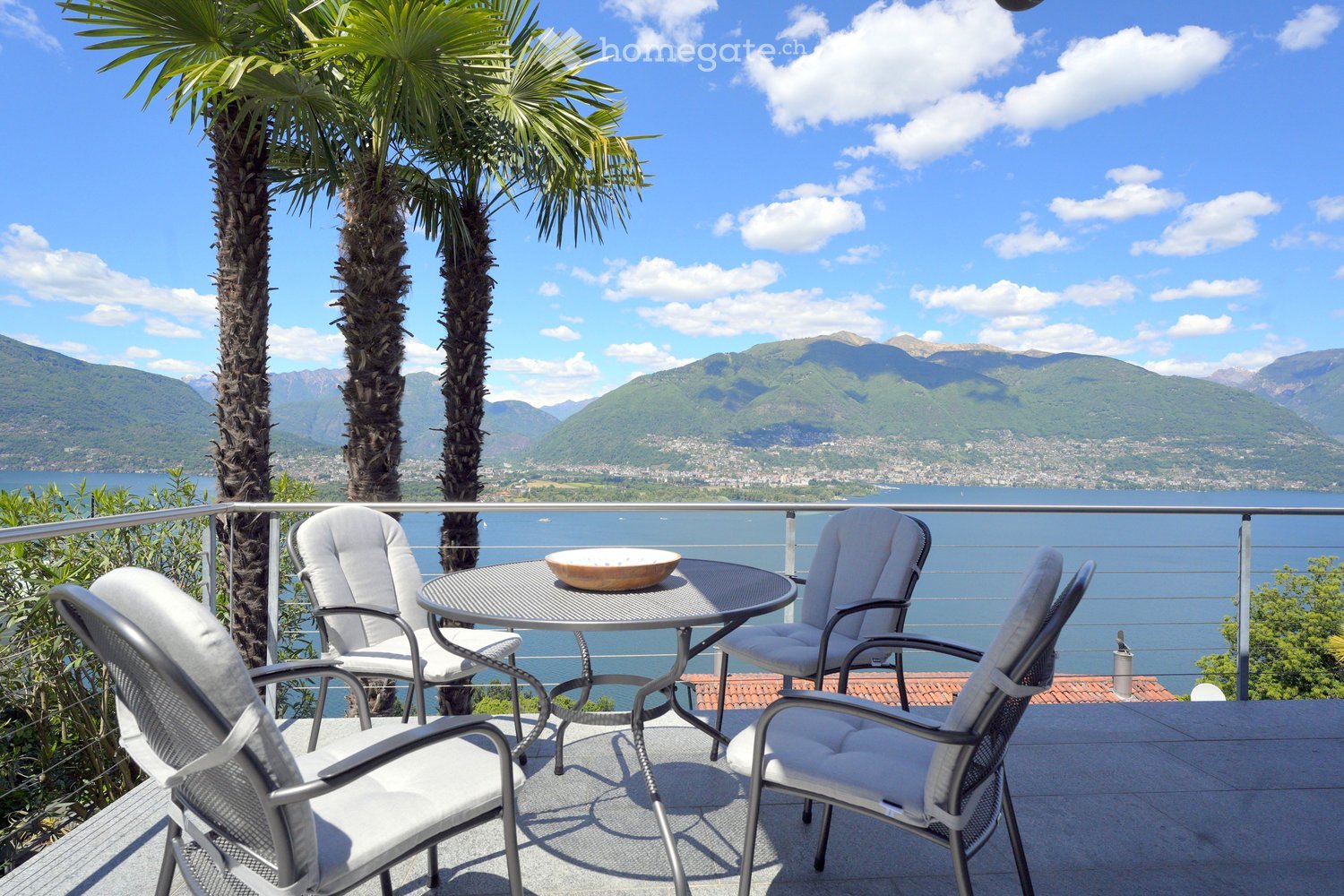 Balcony with a round table, four chairs, and a decorative bowl, overlooking a scenic lake and mountain view with palm trees nearby.