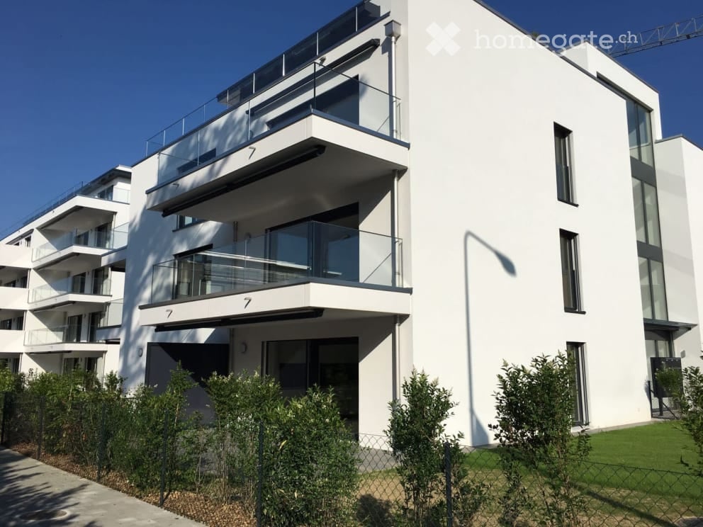 Modern white apartment building with multiple balconies and large windows, surrounded by greenery.