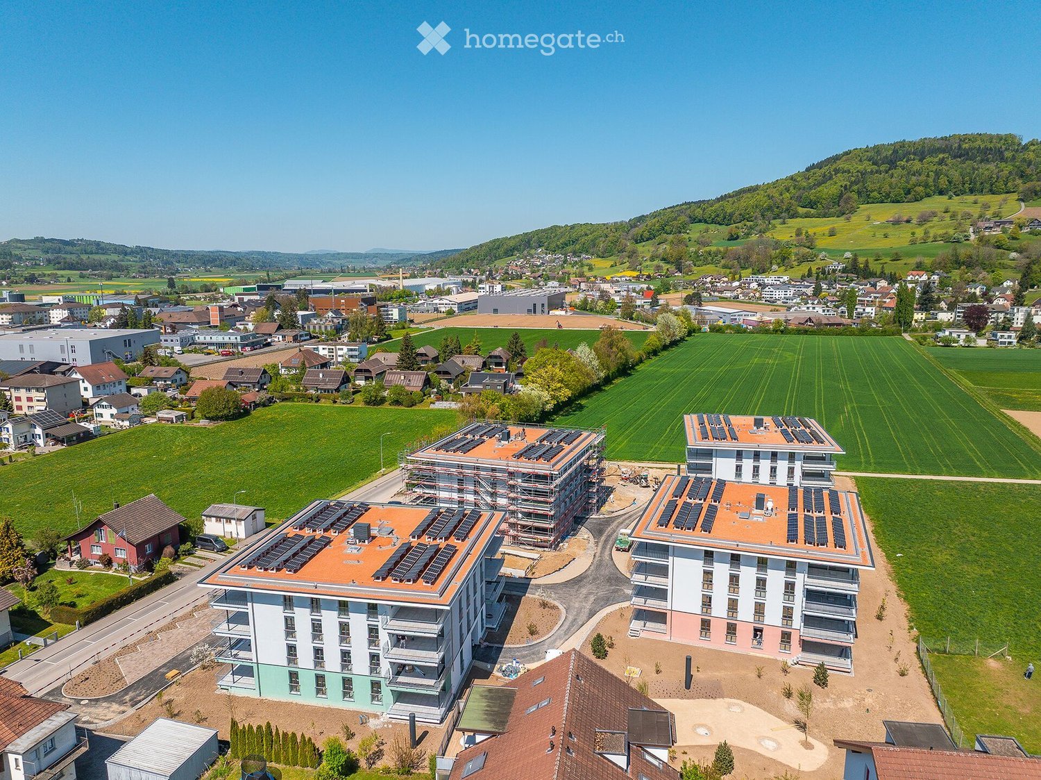 Aerial view of a residential area with modern apartment buildings featuring solar panels on the roofs, surrounded by green fields and scattered houses.