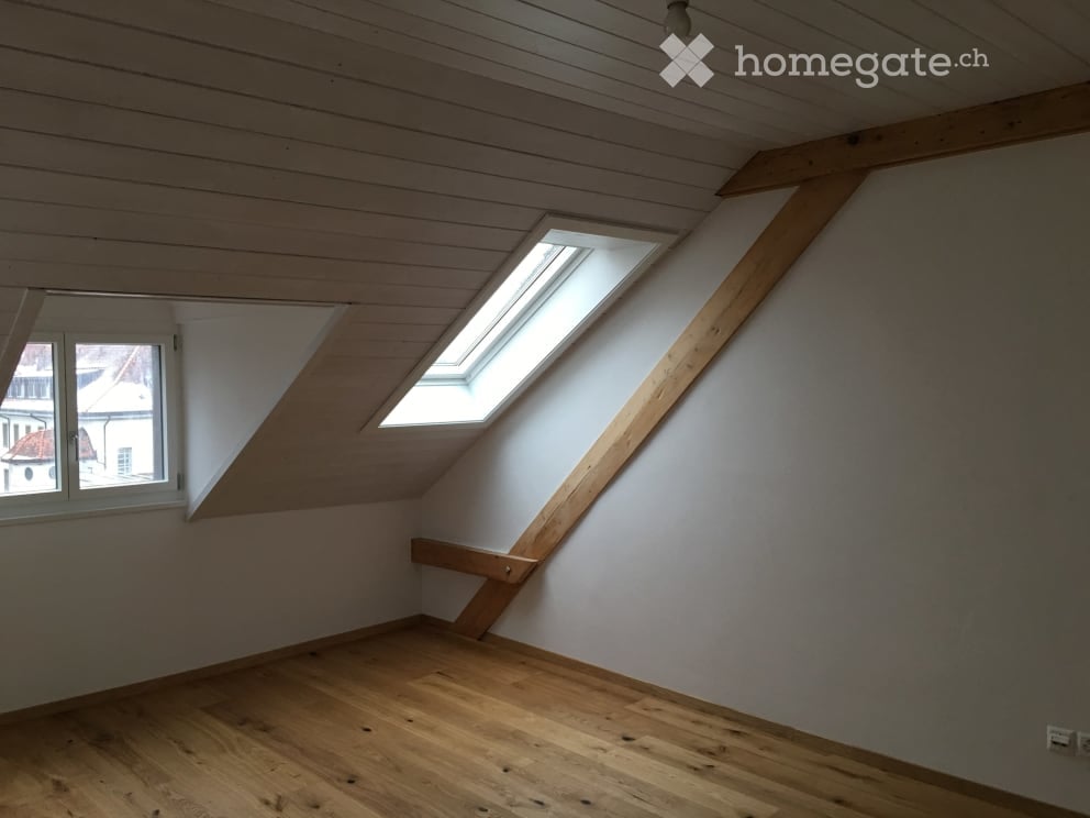 Empty attic room with wooden floor, white walls, exposed wooden beams, and skylight windows.