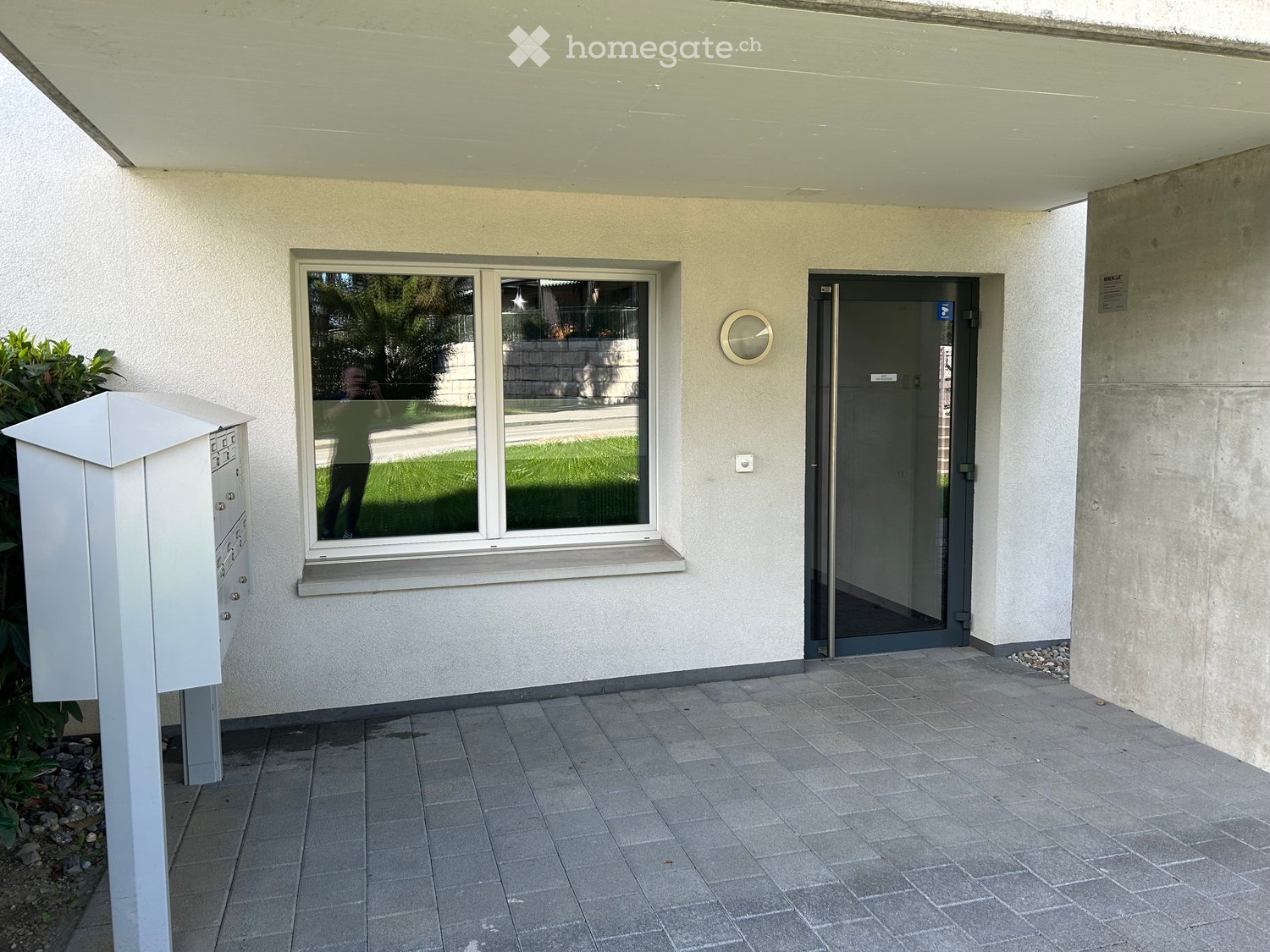 Entrance area of a building with a tiled floor, a window showing a green lawn, a mail box, and a glass door.