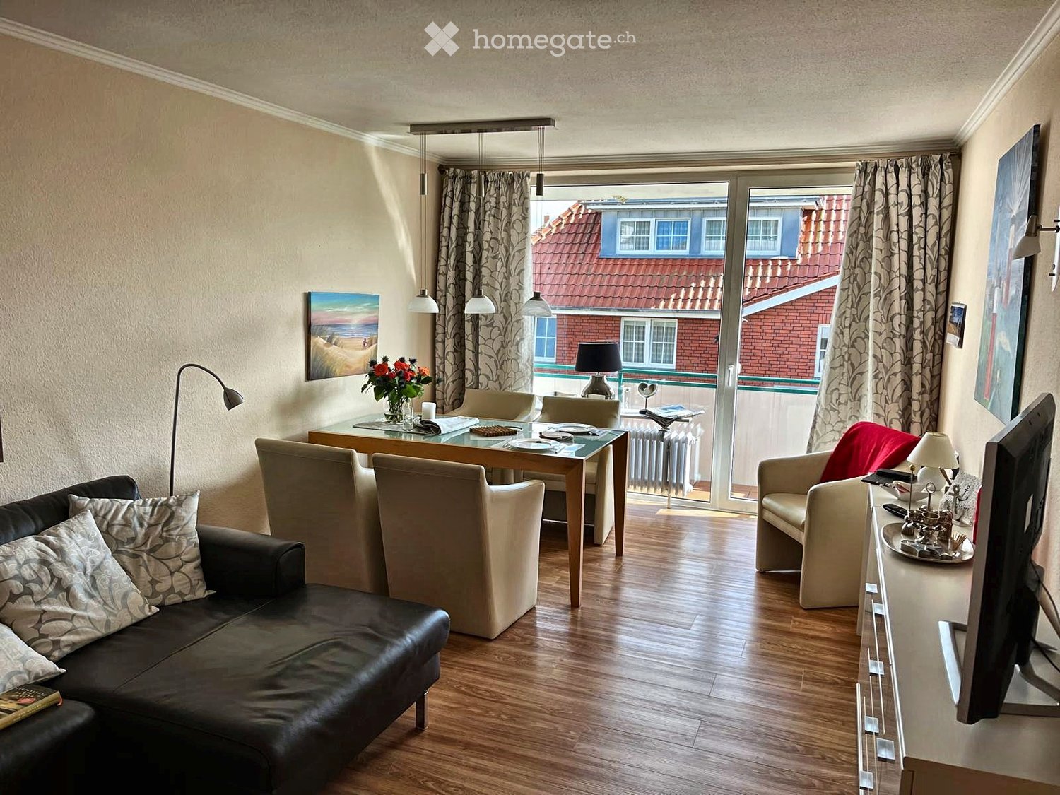 Modern living room with wooden flooring, a black leather sofa, a glass dining table, and a comfortable armchair. The room features large windows with curtains, a view of a neighboring building, and decorative elements like a flower vase and picture frames