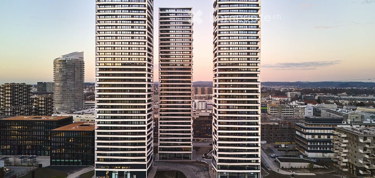 Aerial view of a modern urban area featuring several tall residential skyscrapers with numerous windows, surrounded by other buildings and open spaces.