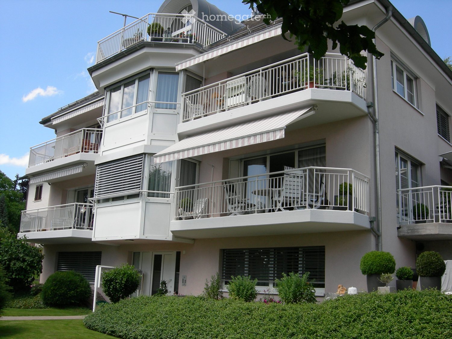 Modern multi-story residential building with multiple balconies, white railings, and outdoor furniture on the balconies. The building has large windows and a well-maintained garden in front.