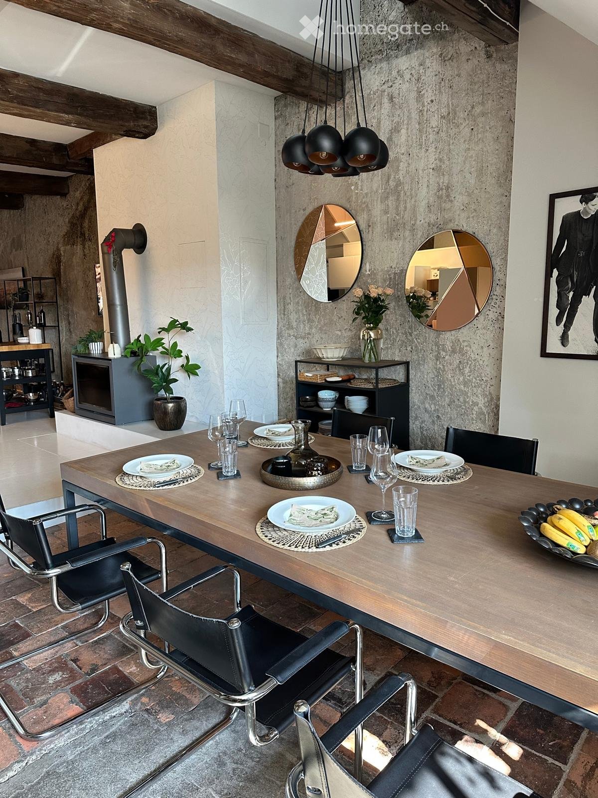 Modern dining area with a long wooden table, black leather chairs, decorative mirrors, a vase with flowers, and a fireplace in the background.