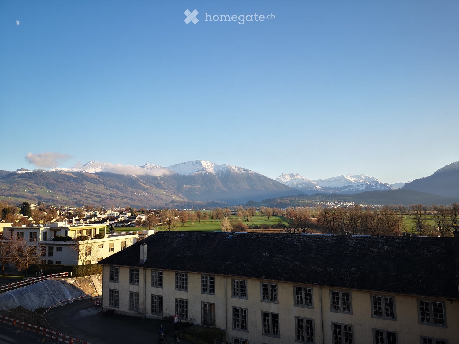 View of a multi-story building with a dark roof and multiple windows, surrounded by a town with visible mountains in the background.